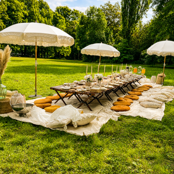Lange Picknicktafel im Park mit Sitzkissen und weißen Sonnenschirmen